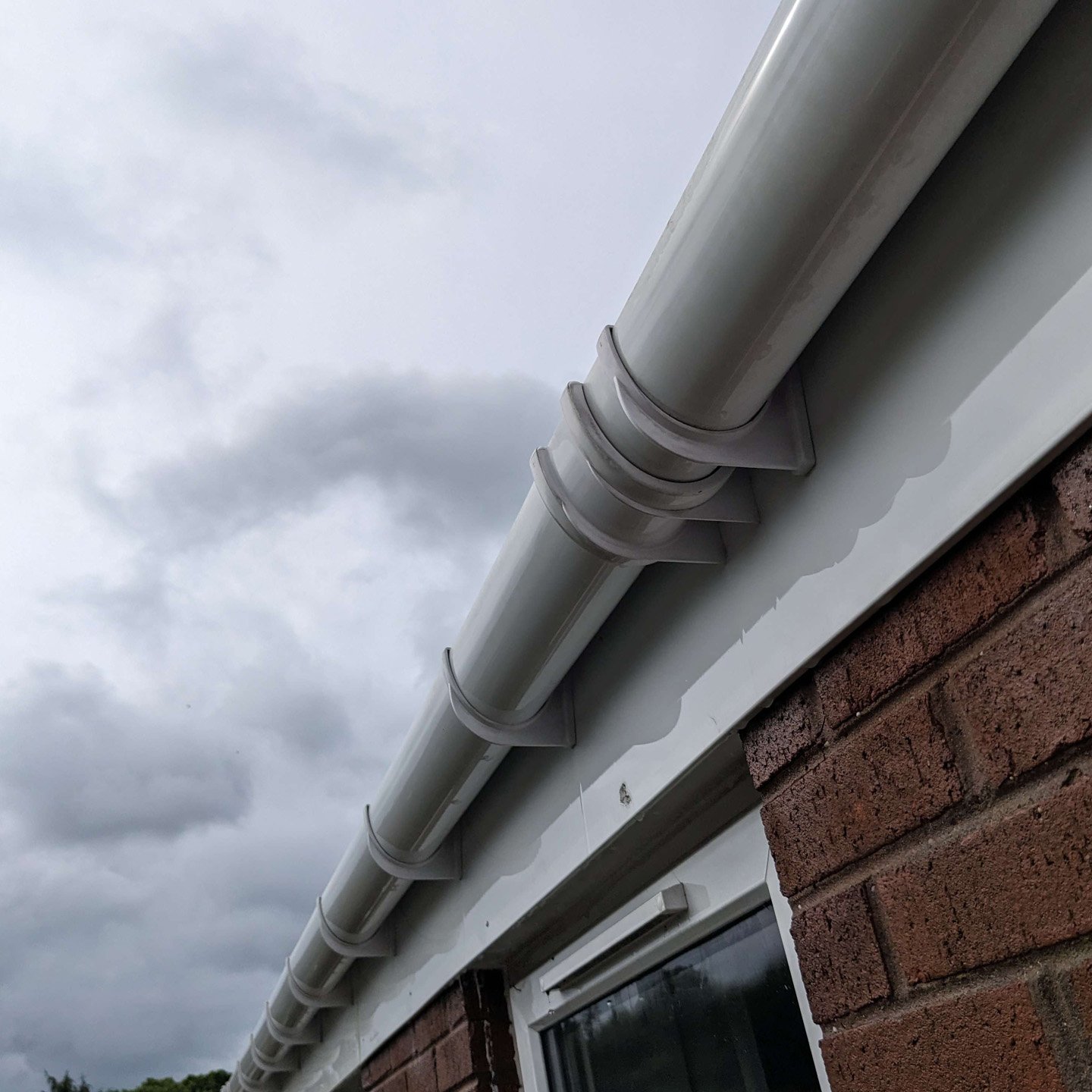 Close-up of a white gutter with leaf guards attached, mounted on a brick house exterior, under a cloudy sky.