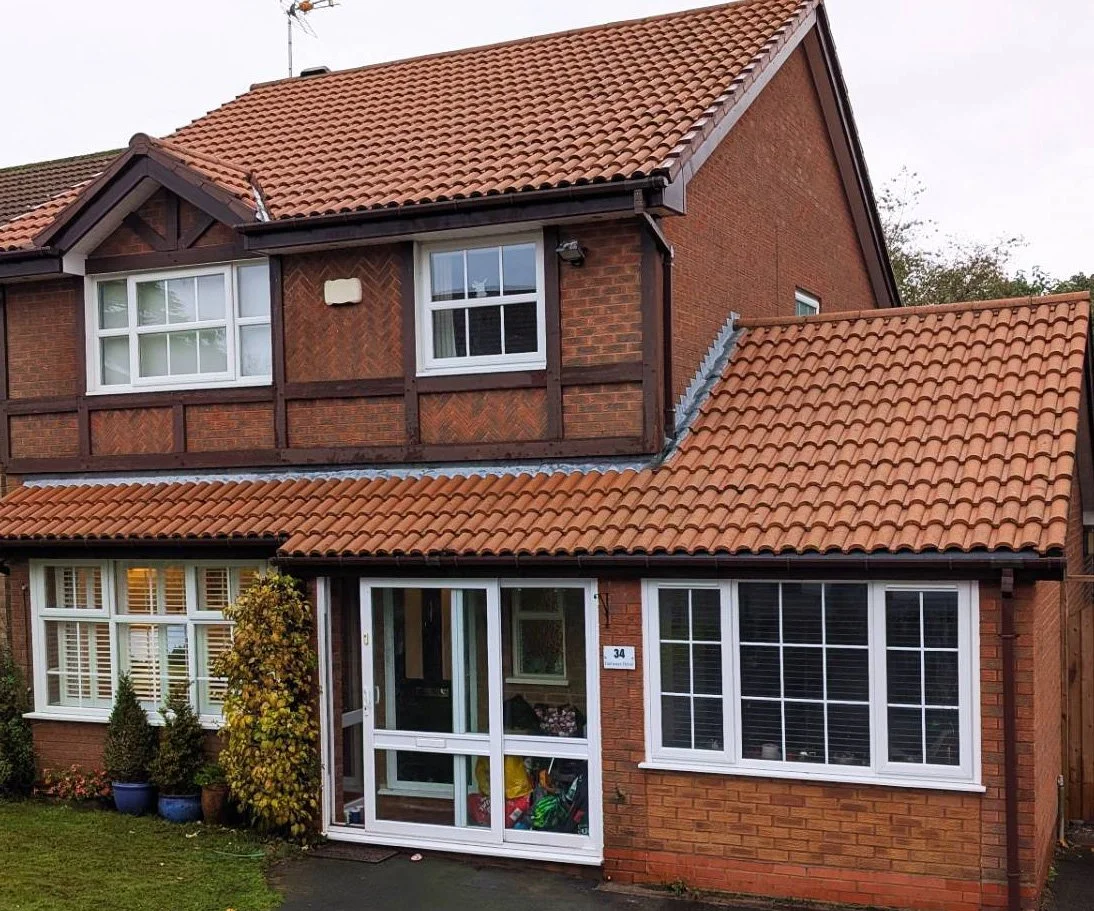 A two-story house with red brick walls and orange tiled roofs, featuring white-framed windows and a front porch with potted plants.