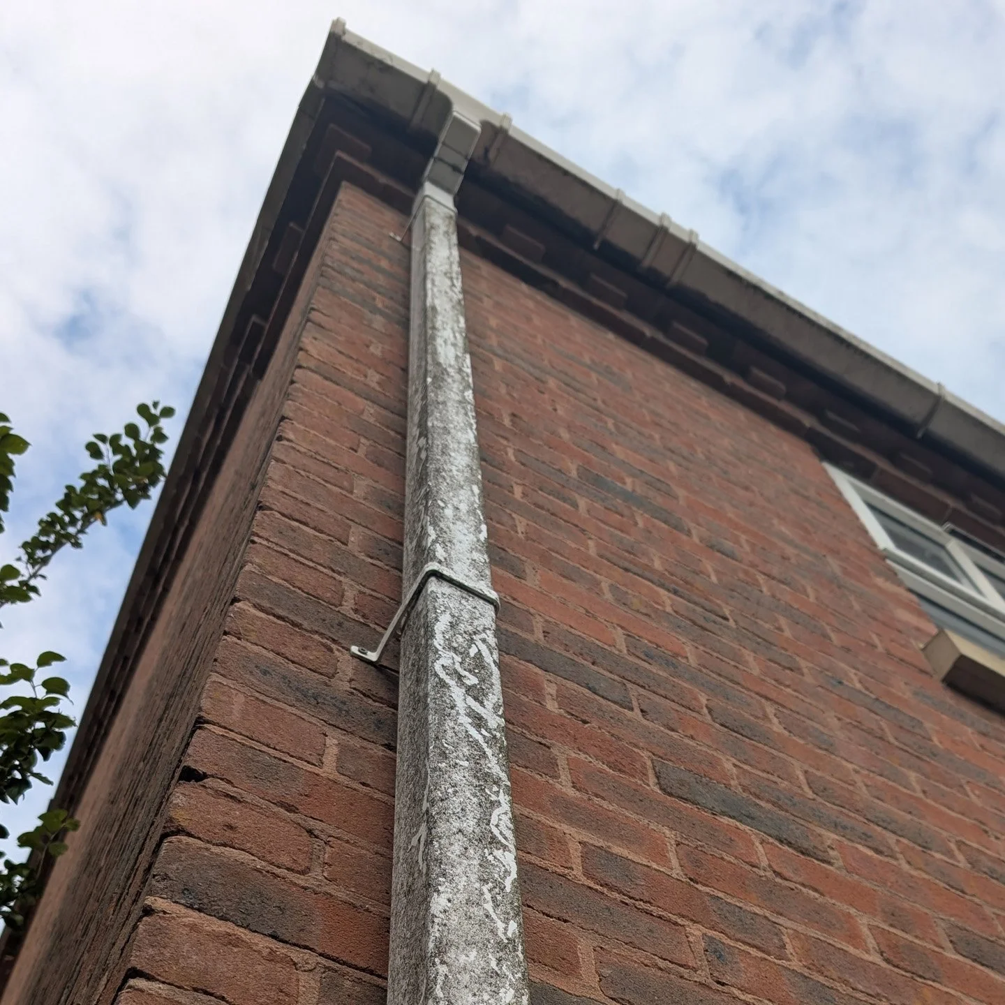 A close-up view of a brick house with a weathered metal downspout and gutter system, taken from ground level looking up at the corner of the building, against a background of a partly cloudy sky.