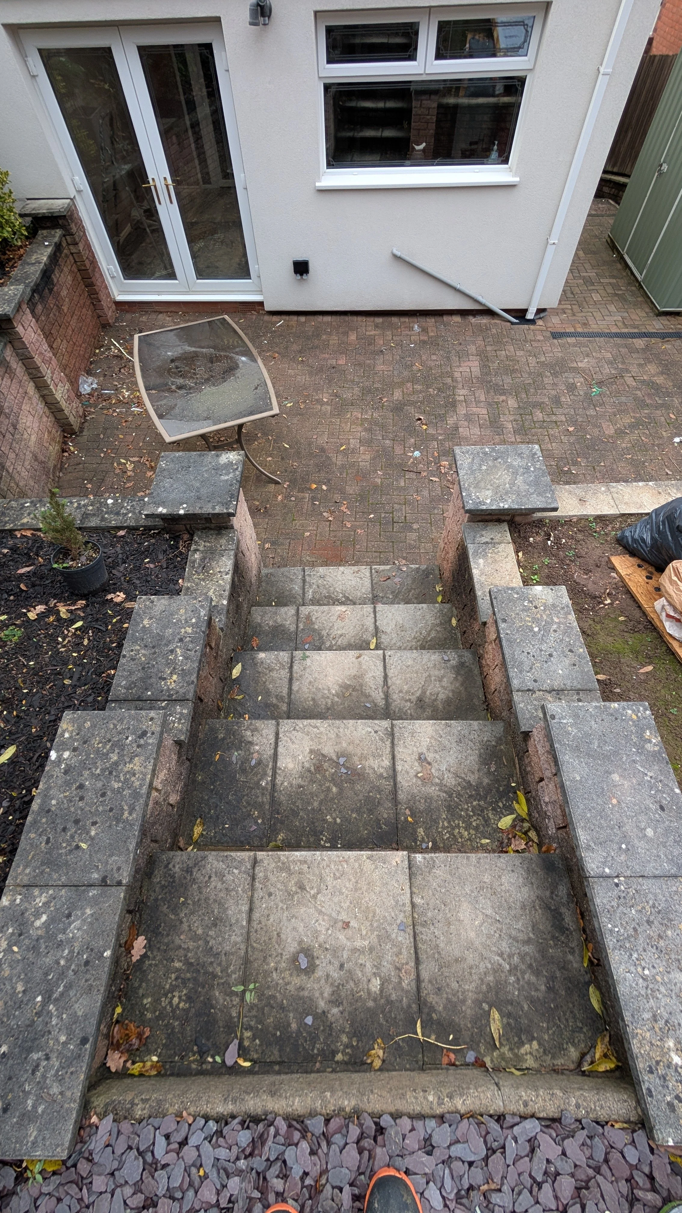View of outdoor stairs made of stone blocks leading down to a paved backyard area with house exterior, glass doors, and windows.