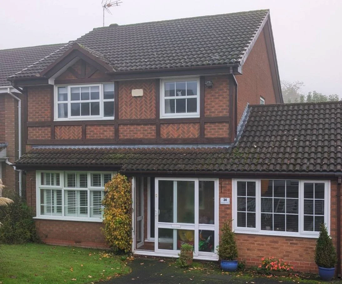 A two-story brick house with a tiled roof and large front windows, surrounded by a small lawn with potted plants and autumn foliage.