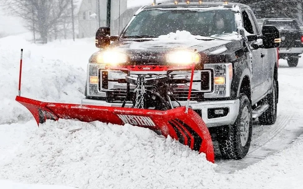 Black pickup truck with snow on hood and windshield, attached to a red snow plow blade clearing snow from the road during a snowstorm.
