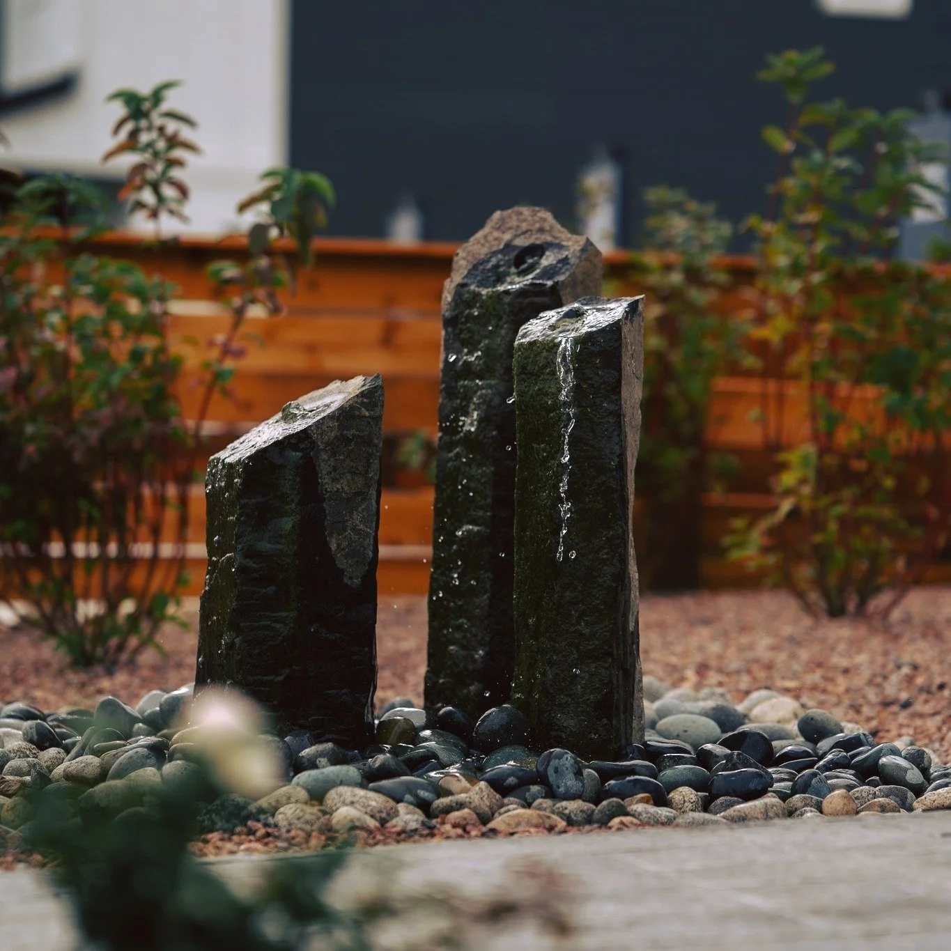 Three black stone water feature with water flowing down their surfaces, surrounded by small pebbles in a landscaped garden with plants and a wooden fence.