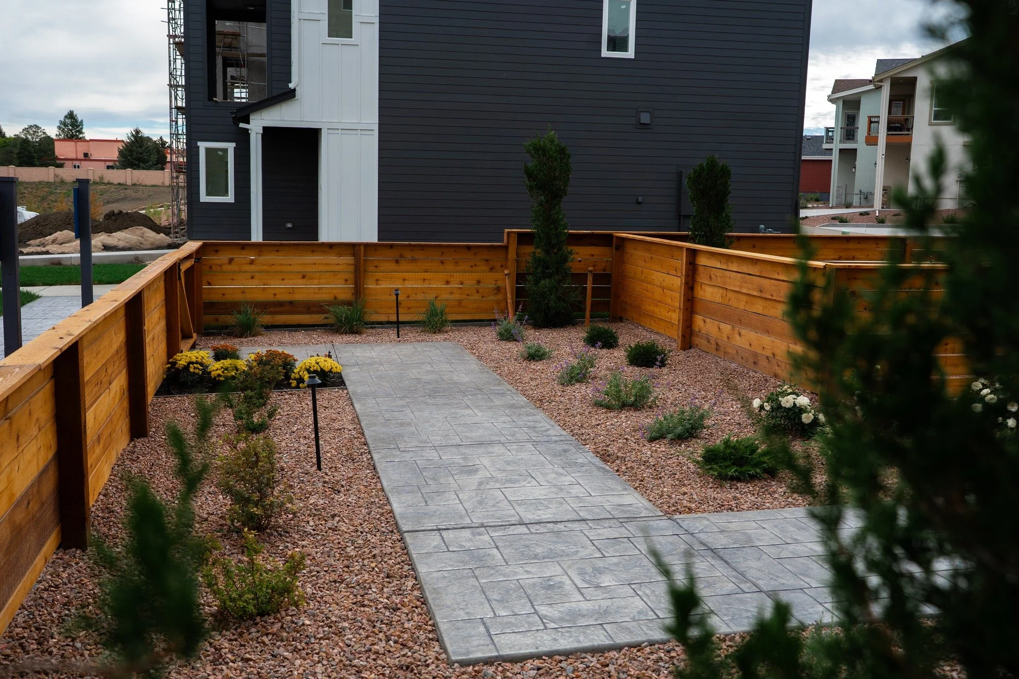 A landscaped backyard with a stone walkway, small plants, shrubs, and a wooden fence, adjacent to a black and white house in a suburban neighborhood.