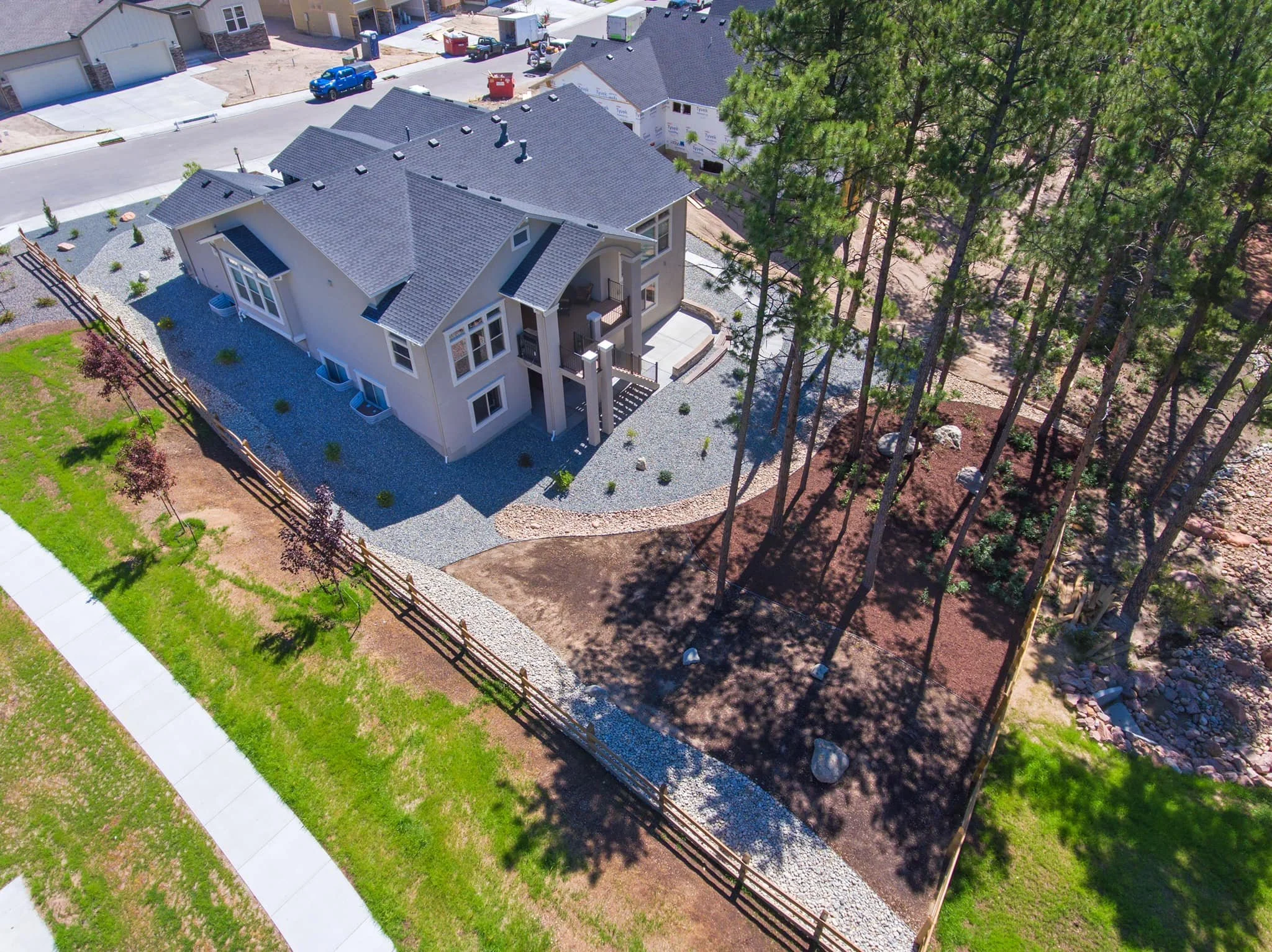 Aerial view of a modern two-story house with a gray roof, surrounded by gravel and landscaped yard, with neighboring houses and a street in the background. Tall trees are on one side of the yard.