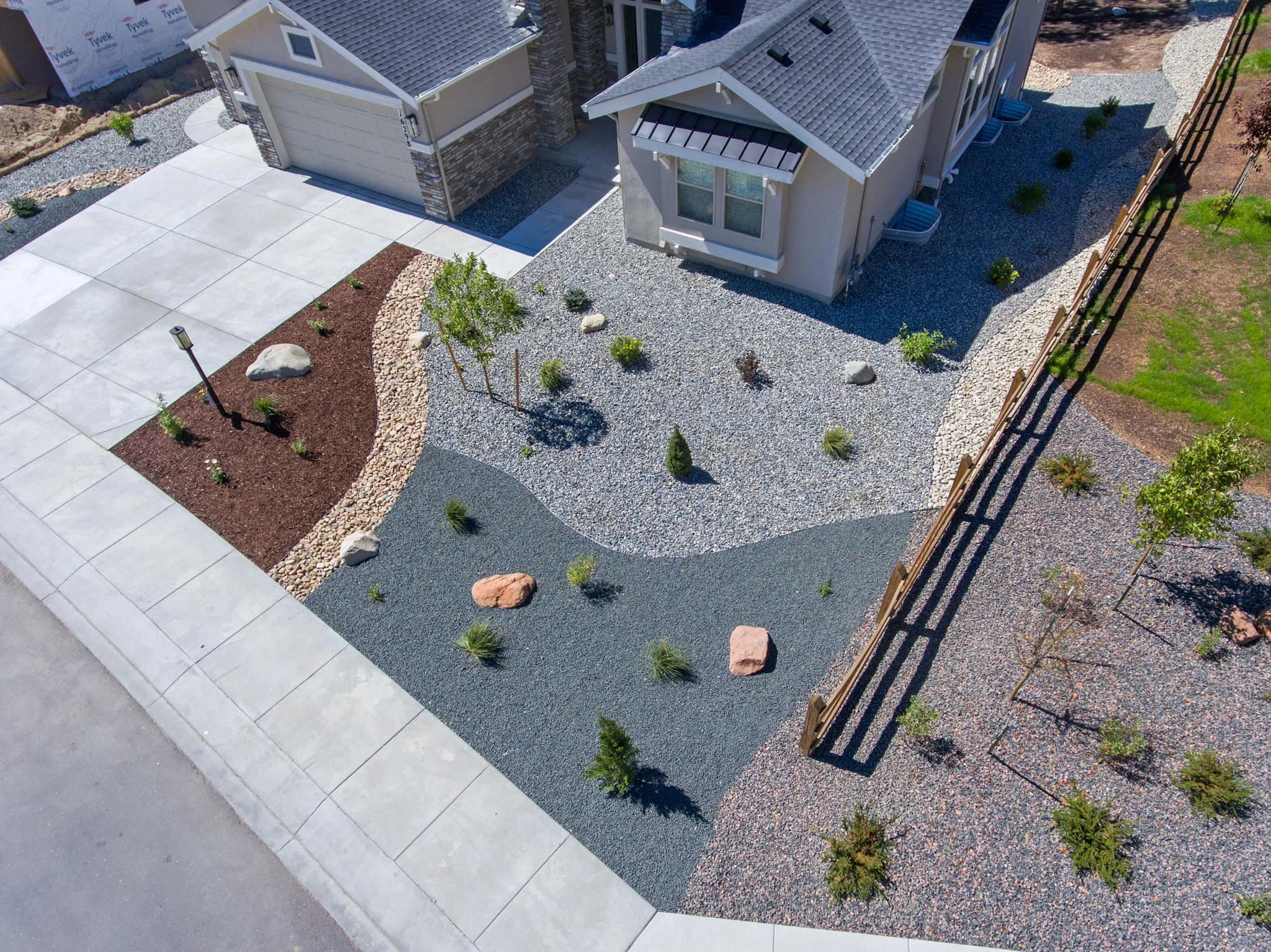 An aerial view of a landscaped yard with different colored gravel sections, small plants, rocks, a wooden fence, a lamppost, and a house with a concrete driveway.