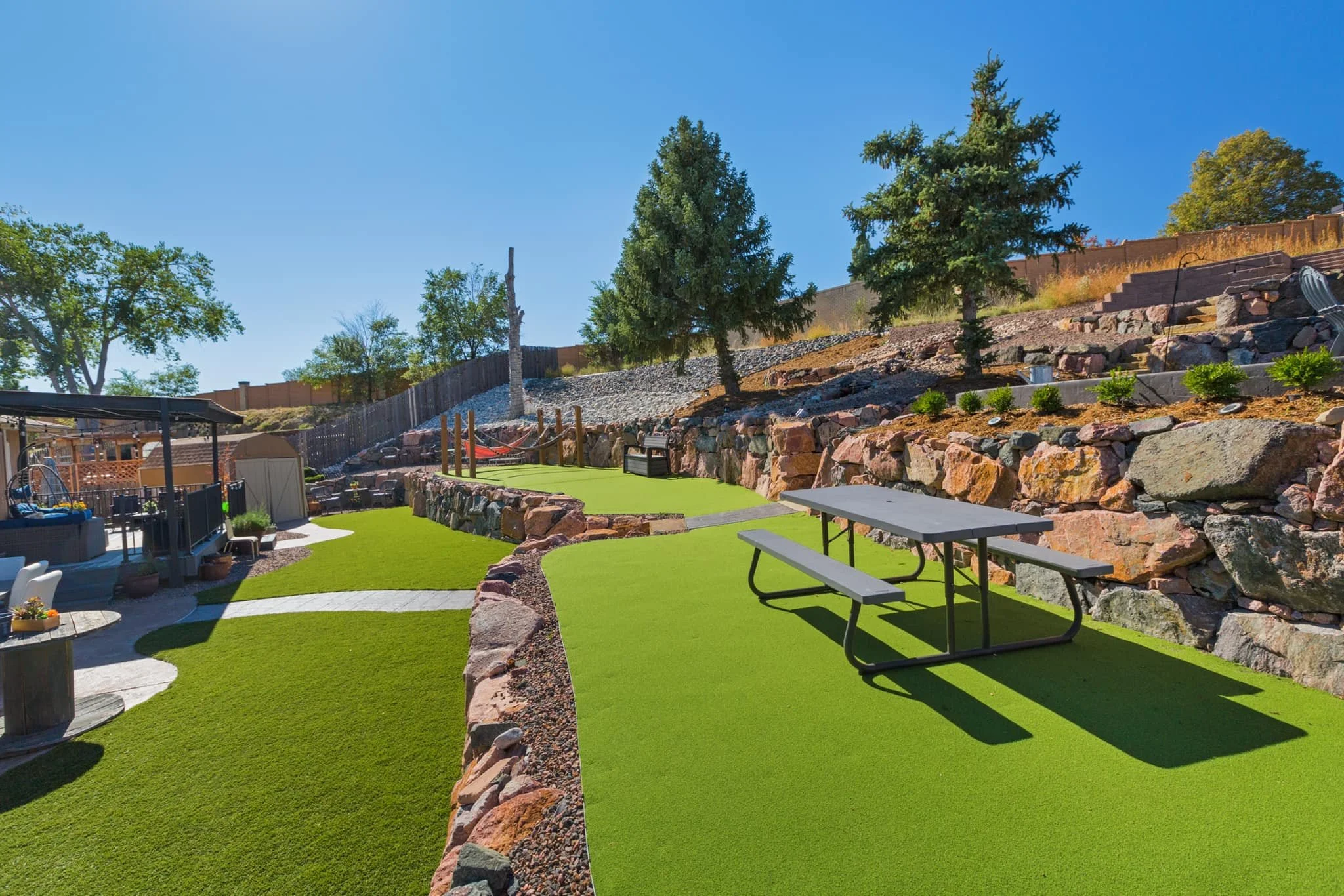 Backyard with artificial grass, stone retaining wall, trees, picnic table, and outdoor seating area on a sunny day.