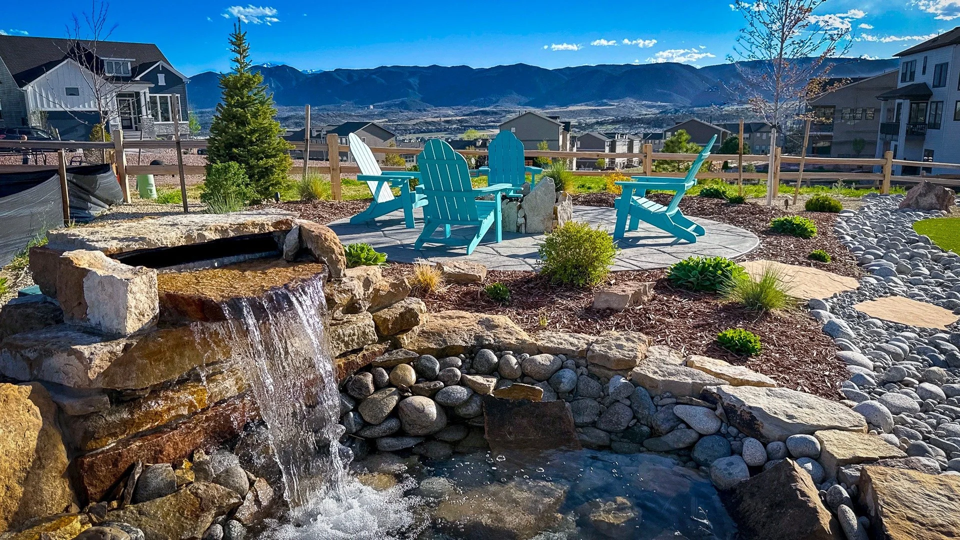 A landscaped backyard with a small waterfall, a circular stone patio with four turquoise Adirondack chairs, surrounded by plants, rocks, and a mountainous background.