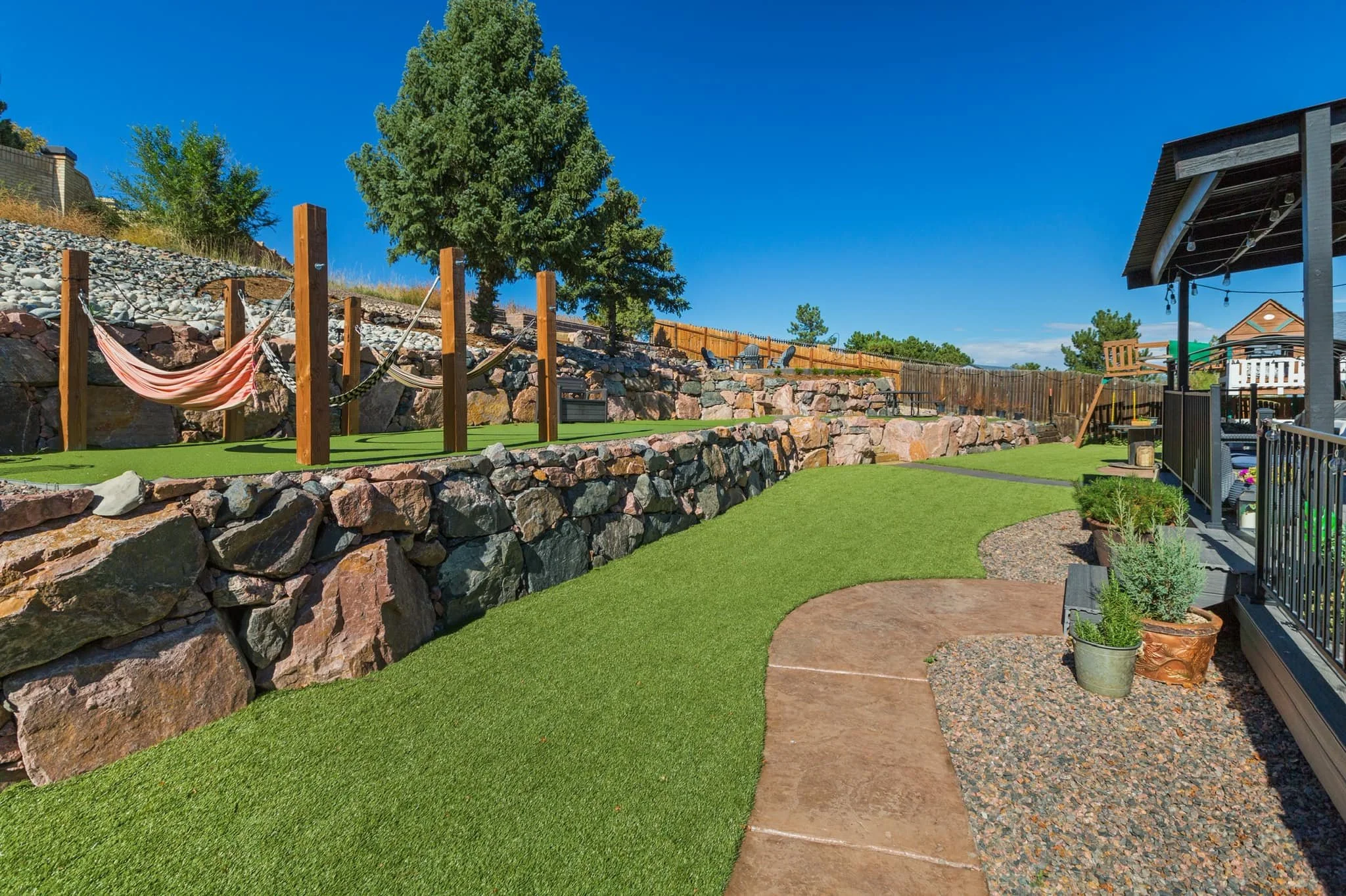Backyard with artificial grass, stone wall, and outdoor seating area under a covered porch with potted plants.