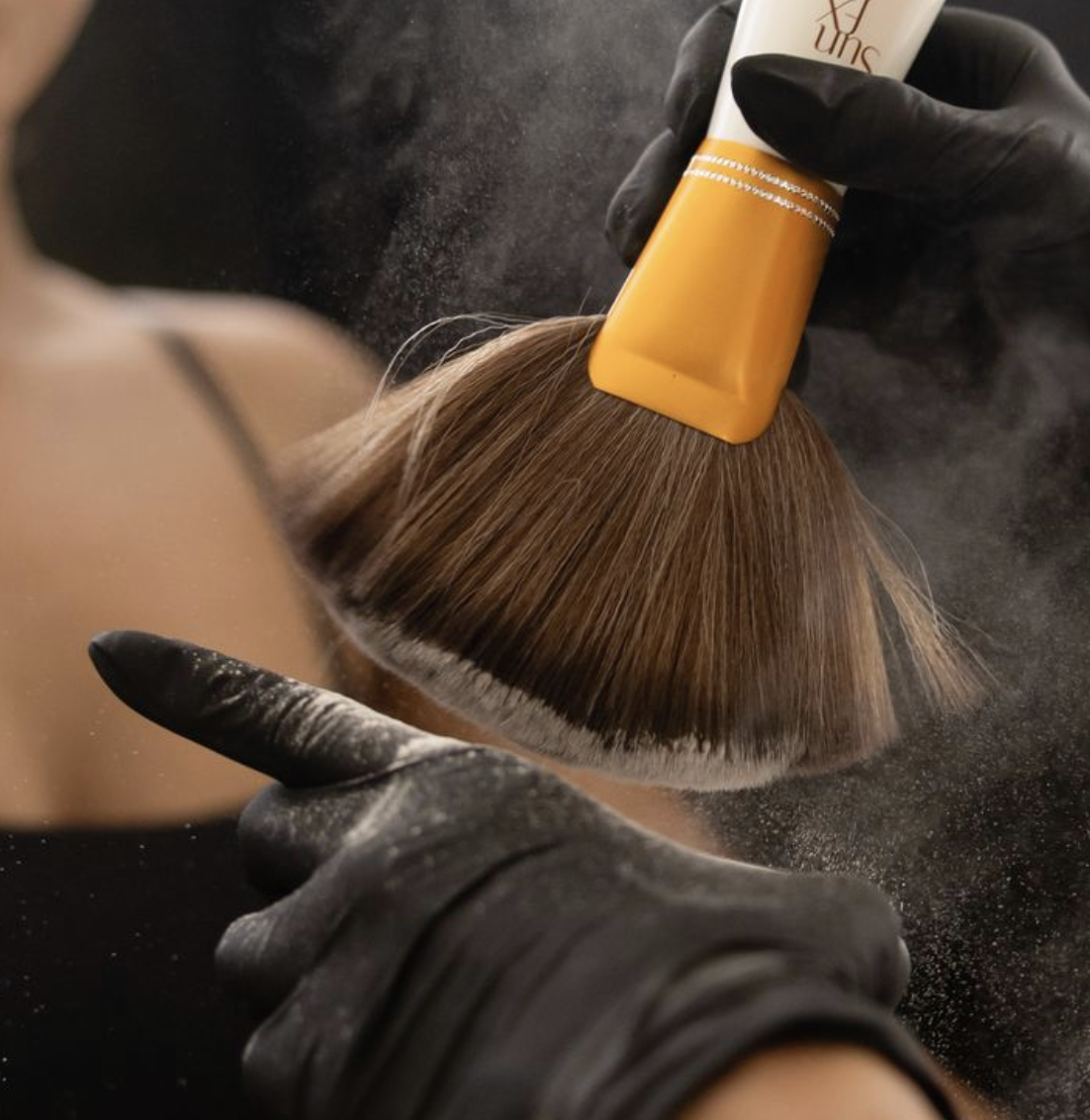 Person applying powder to a makeup brush with a yellow handle using gloved hands.