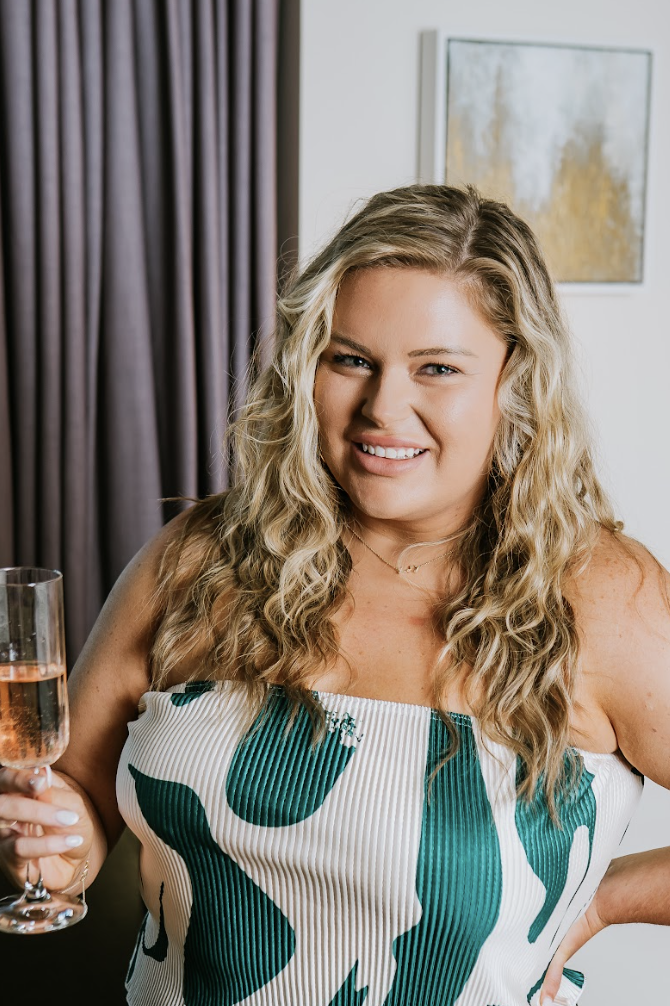 A woman with long wavy blonde hair smiling and holding a glass of rosé wine.