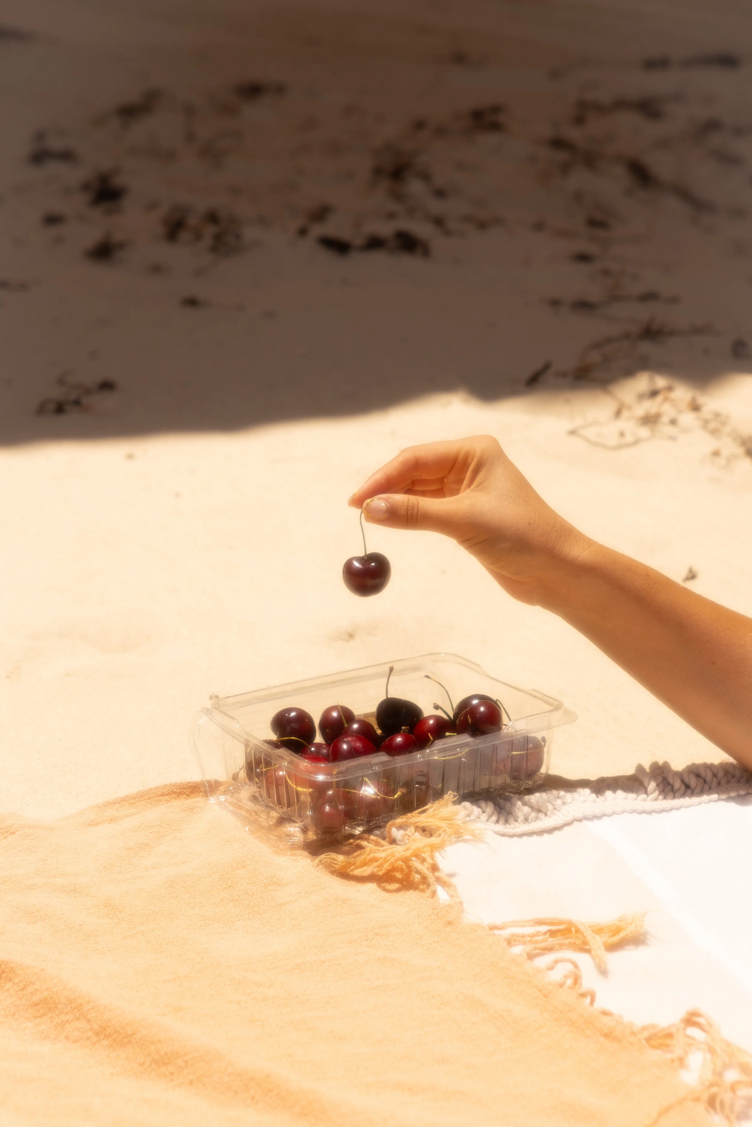 A hand picking up a cherry from a plastic container filled with cherries on a sandy beach.