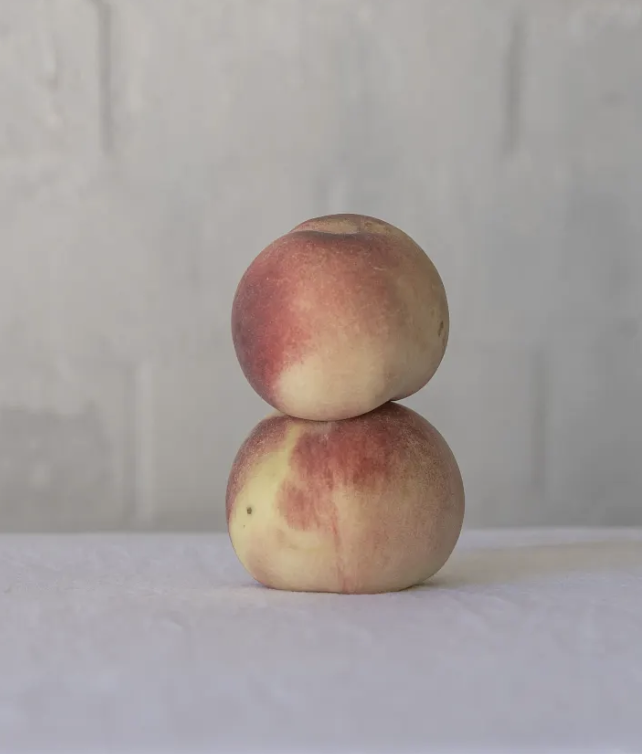 Two peaches stacked on top of each other on a white surface with a plain gray wall in the background.