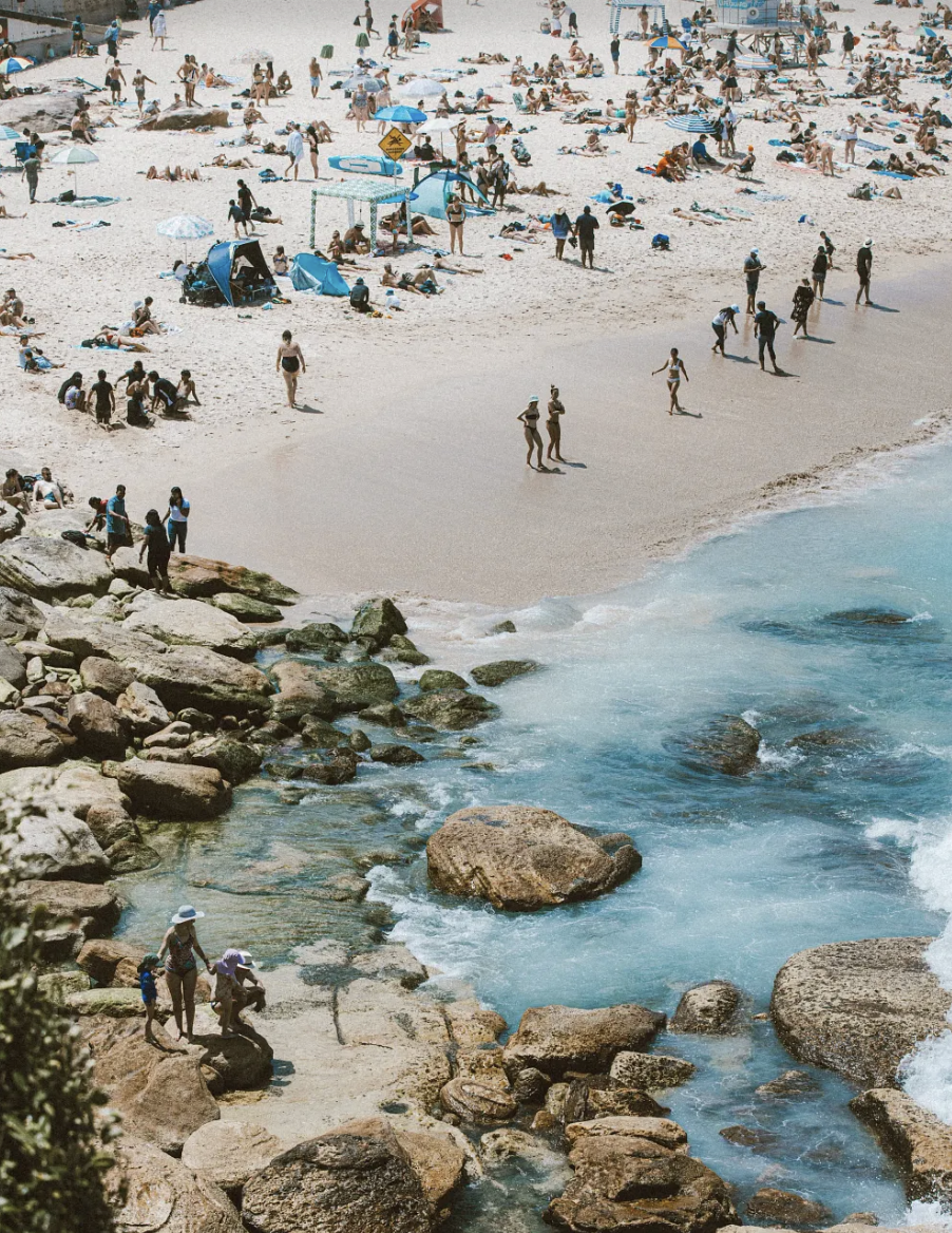 A crowded beach with many people sunbathing, swimming, and walking along the shoreline. There are umbrellas, tents, and beach equipment scattered across the sand, with rocky areas near the water.