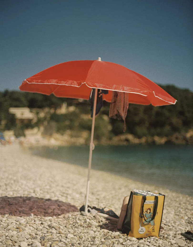 A beach scene with a red beach umbrella, a bag, and a towel on a pebbled shore, with water and distant hills in the background.