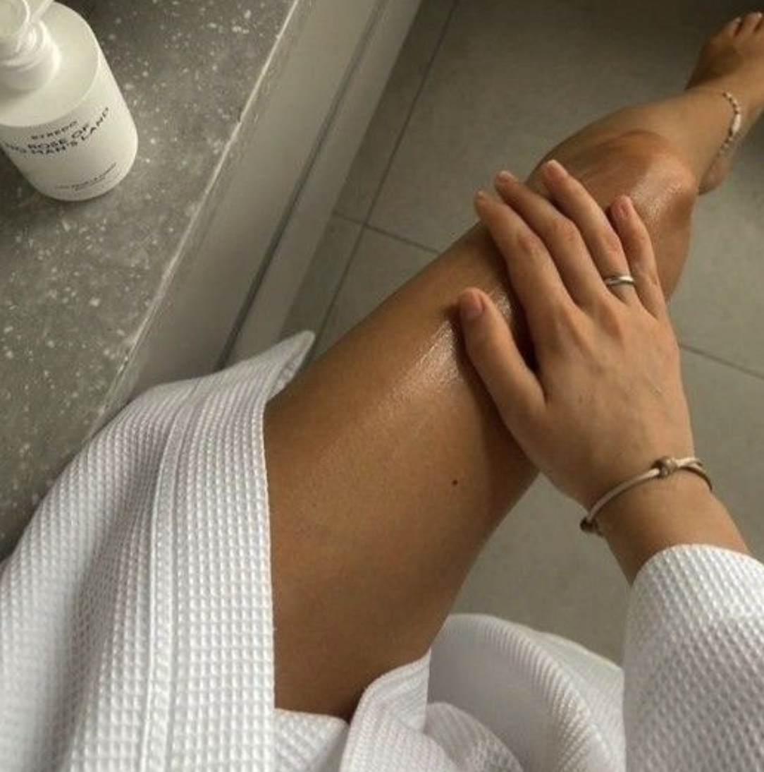 A person applying lotion to their leg while sitting on a tiled bathroom floor, with a bottle of lotion on the counter nearby.