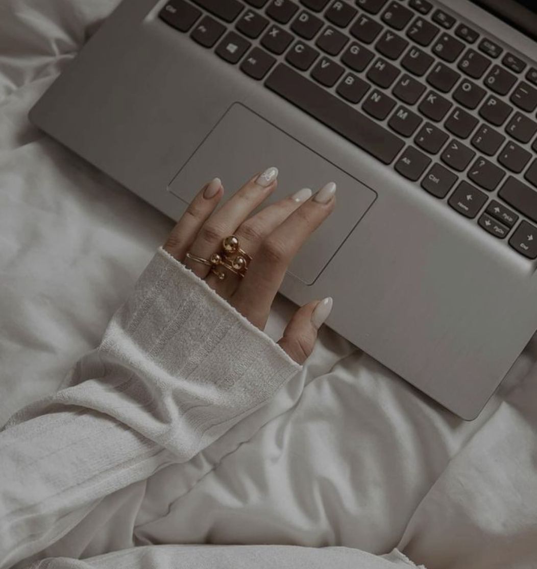 A person's hand with manicured nails and wearing rings, resting on a silver laptop keyboard, on a white bed or blanket.