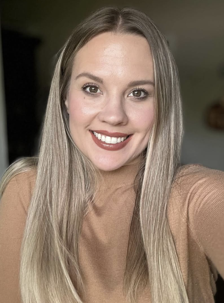 A young woman with long, straight blonde hair, smiling, wearing a beige top, indoors.