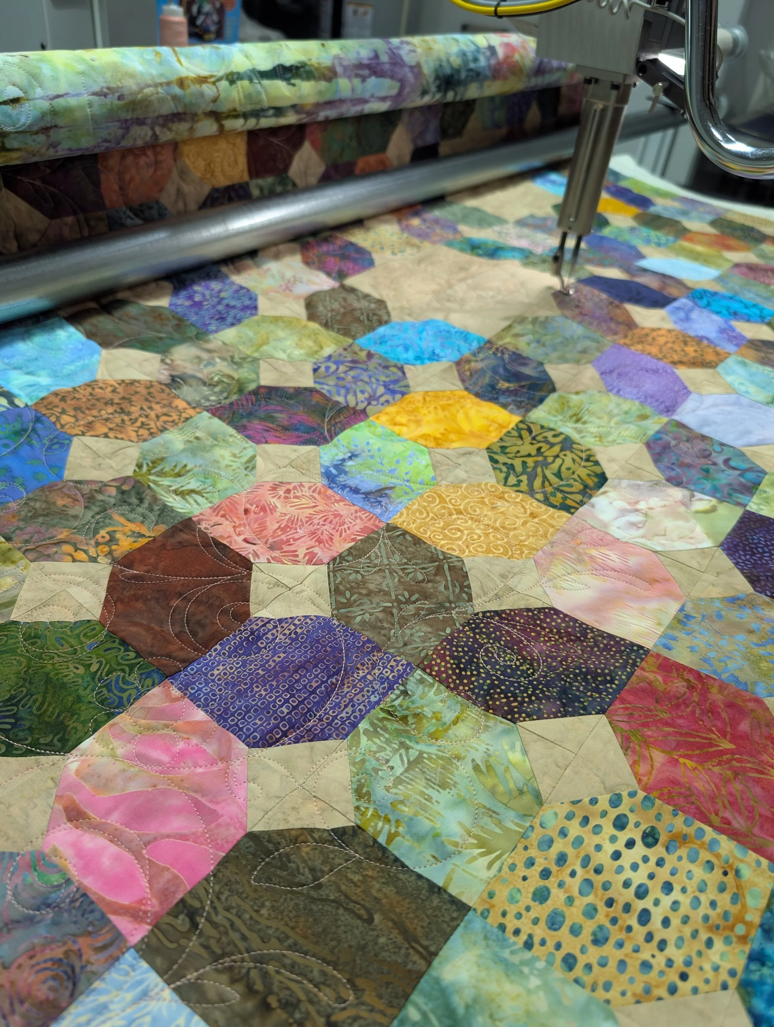 Close-up of a colorful quilt with various patterned fabric patches and quilting stitches, with a quilting machine guiding the fabric.