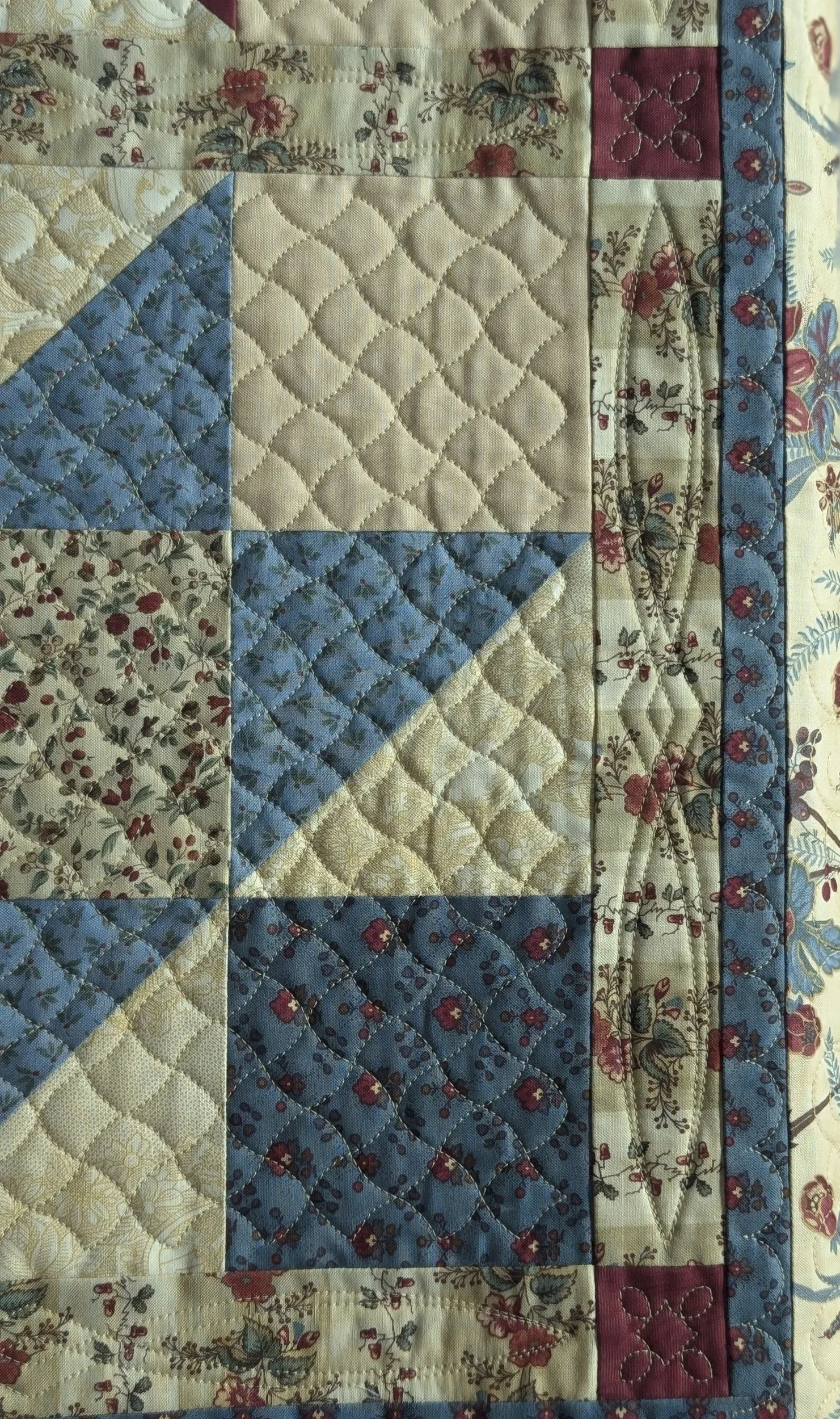 Close-up view of a quilt with floral and patterned fabric pieces sewn together in a patchwork design, featuring beige, blue, red, and cream colors.