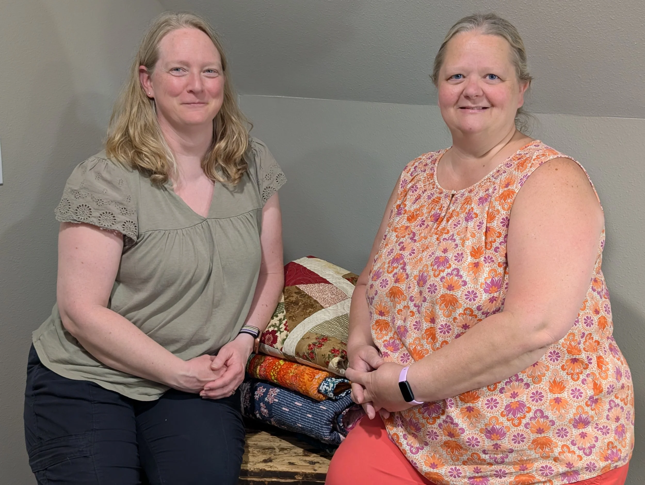 Two women standing side by side inside a room with a neutral-colored wall behind them. The woman on the left has blonde hair, wearing a light green top, and black pants. The woman on the right has gray hair, wearing a sleeveless orange and purple floral dress, and coral-colored pants. They are posing with a quilt on a table in front of them.