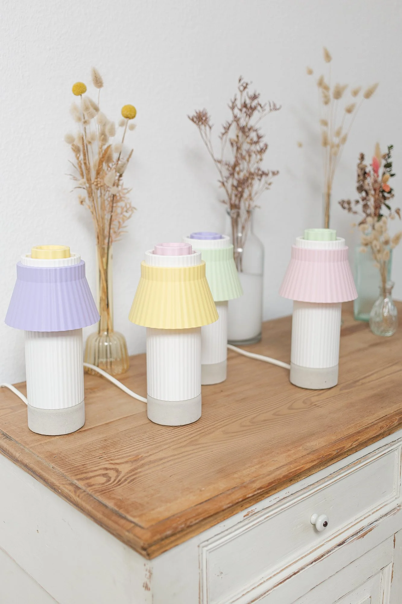 Drei dekorative Tischlampen mit pastellfarbenen Lampenschirmen in lila, gelb und rosa auf einem Holz-Tisch, dahinter Vasen mit getrockneten Blumen.