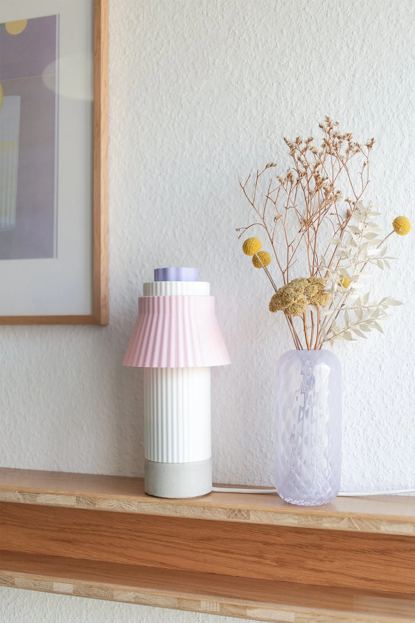 Vase mit getrockneten Blumen und eine designte Lampe auf einem Holzregal an einer weißen Wand.