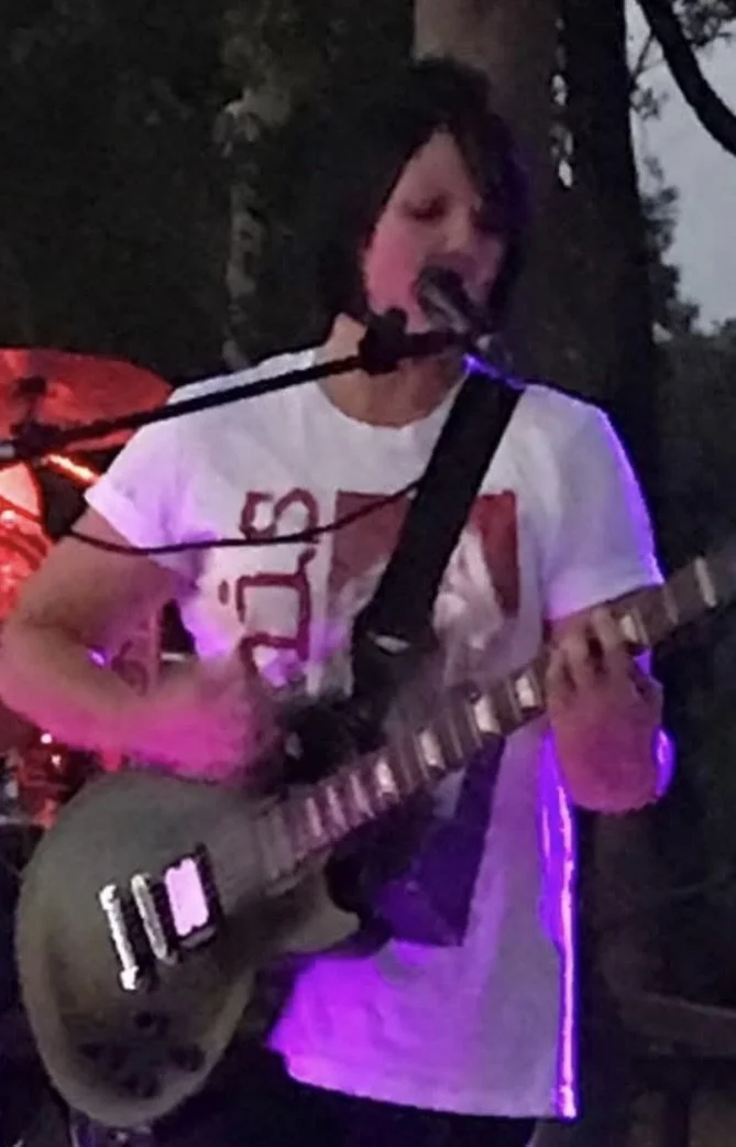 A person playing an electric guitar and singing into a microphone during an outdoor performance at dusk, with trees and purple lighting visible.