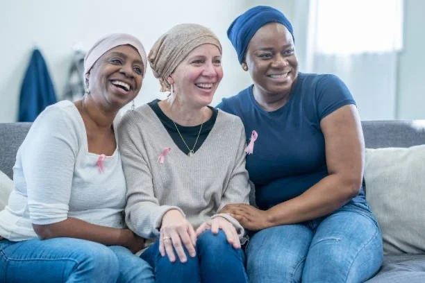 Three women sitting on a couch, smiling, with pink ribbons on their clothing supporting breast cancer awareness.