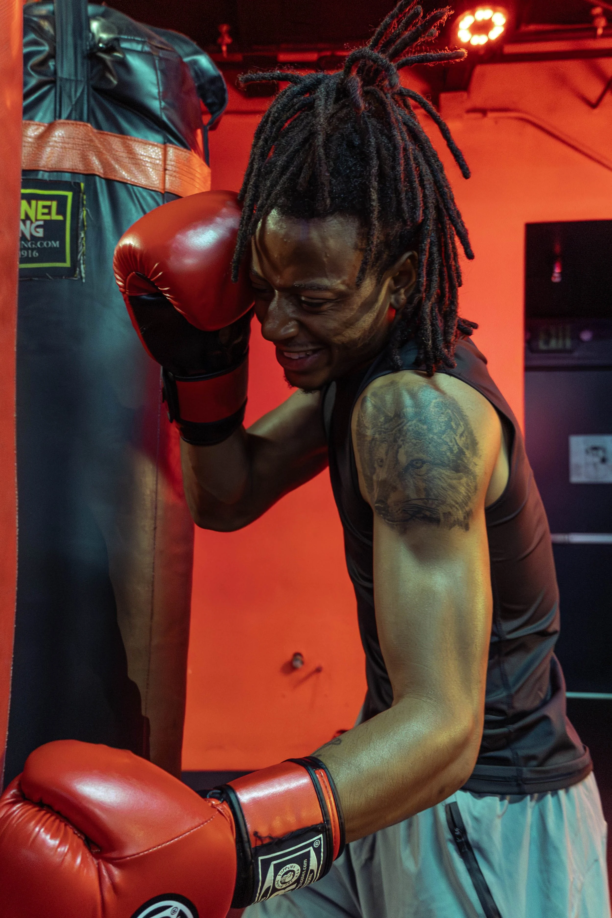 A boxer in gray athletic shorts and black tank top sparring with a punching bag, wearing red boxing gloves, in a gym with red lighting.