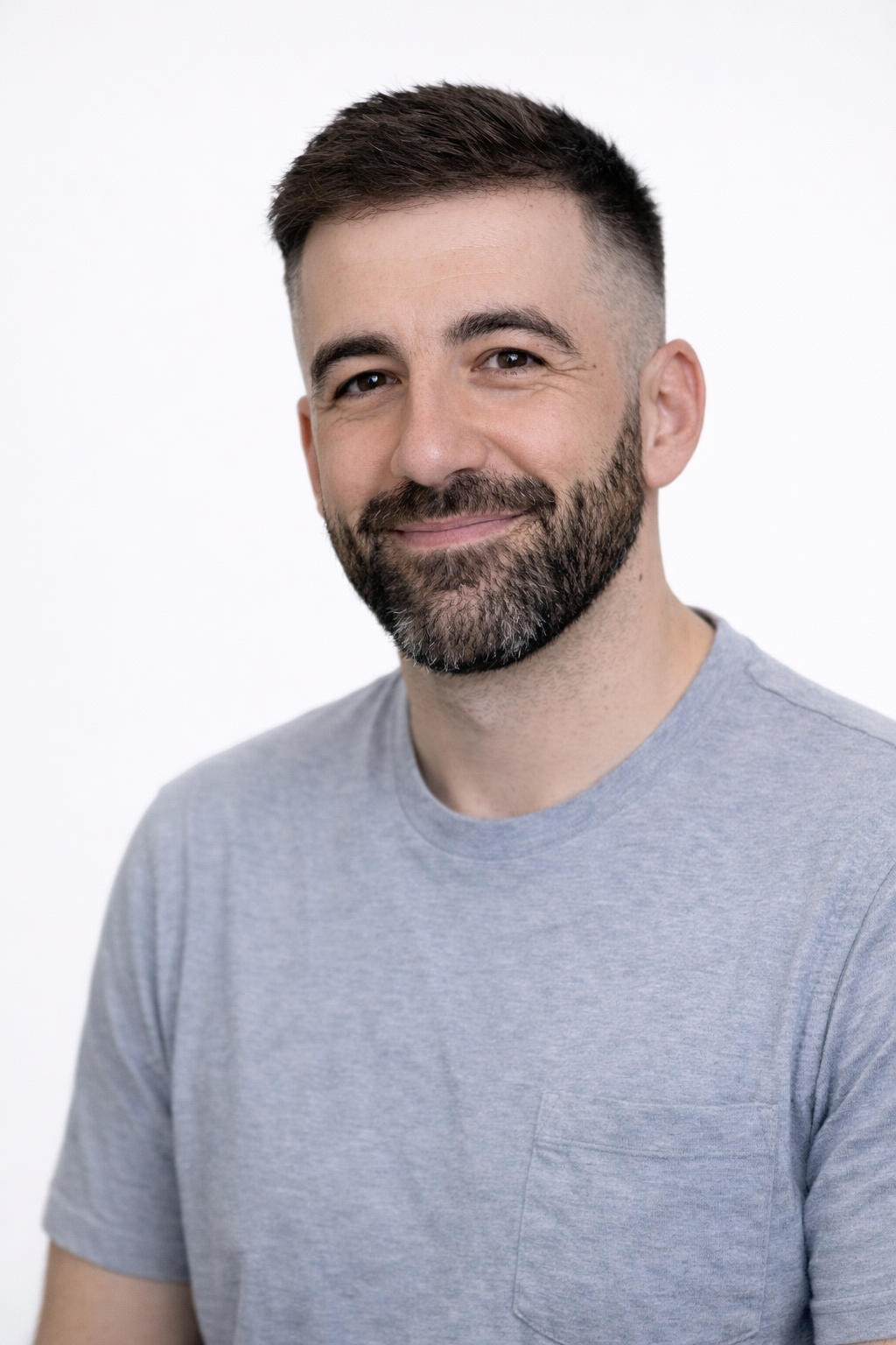 A man with short dark hair and a beard, smiling, wearing a gray t-shirt against a plain white background.