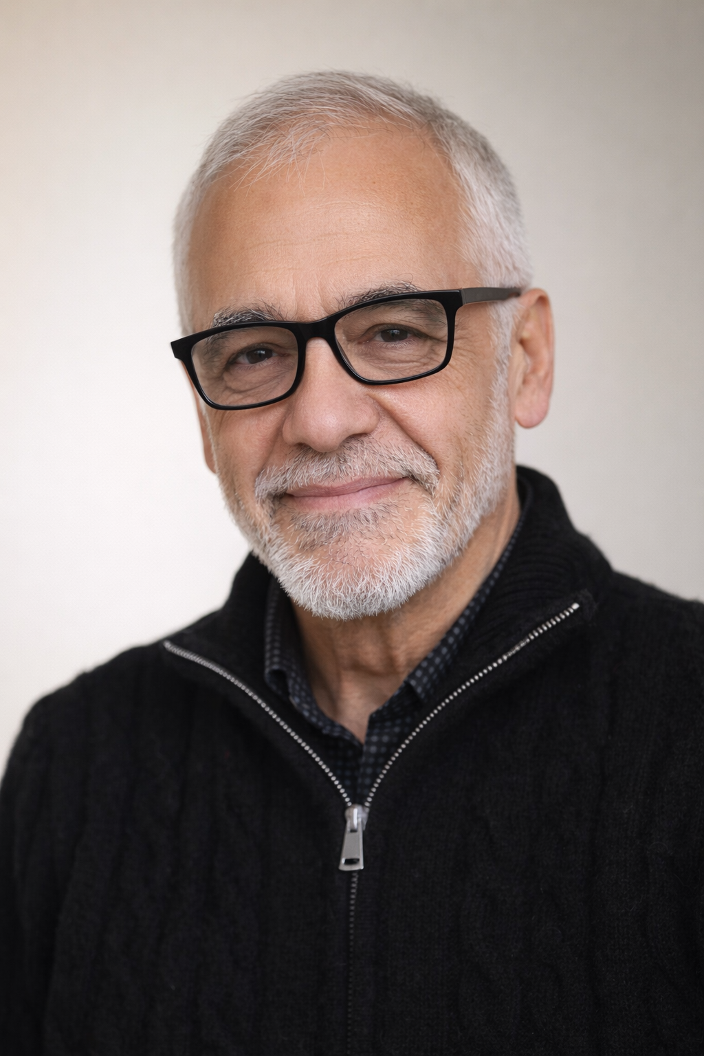 Close-up photo of an older man with white hair, beard, and black glasses, smiling softly, wearing a black zip-up sweater over a checkered shirt.
