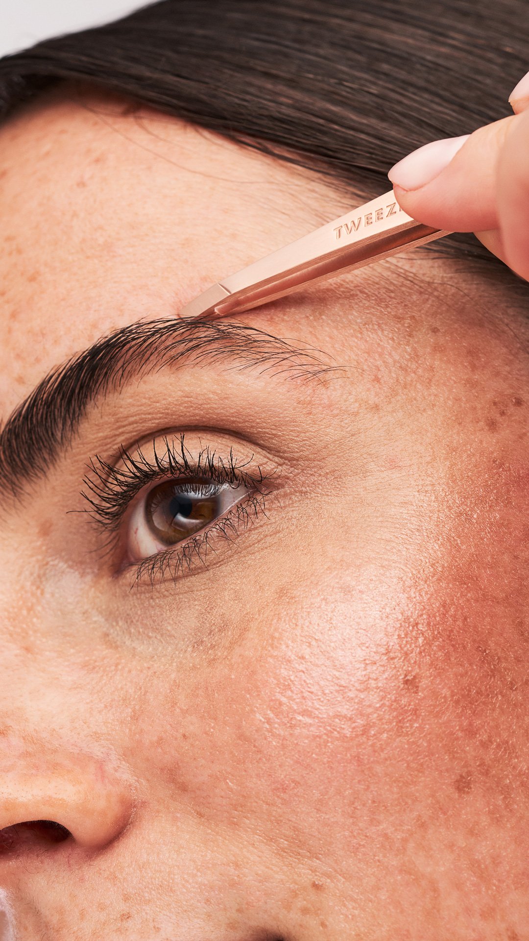 White model, picking at her eye brow with a rose gold Tweezerman tweezers.