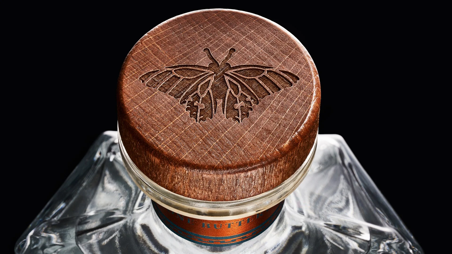 Close-up of a glass bottle with a wooden cap engraved with a butterfly design, against a black background.