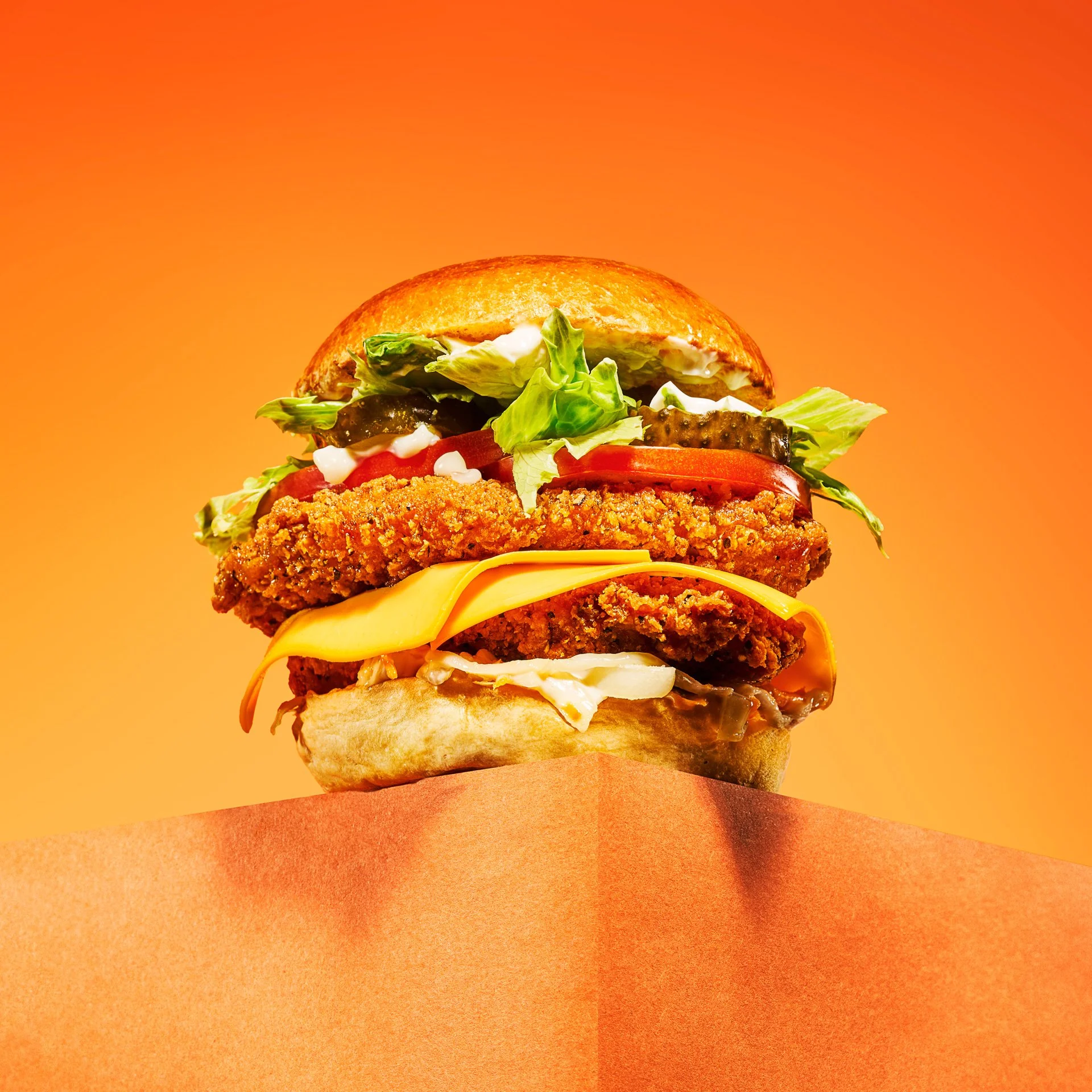 Close-up of a large, layered chicken sandwich with lettuce, tomato, pickles, cheese, fried chicken, and sauce on a bun, set against a gradient orange background.