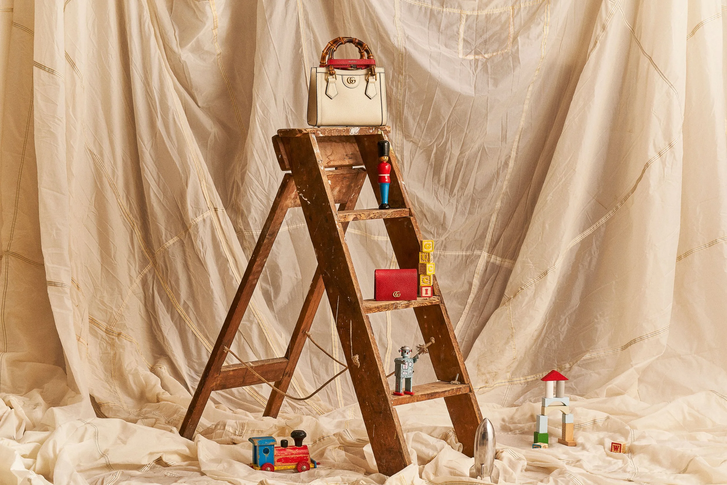 A wooden toy step ladder with various small toys and objects placed on it and around it, against a beige fabric backdrop.