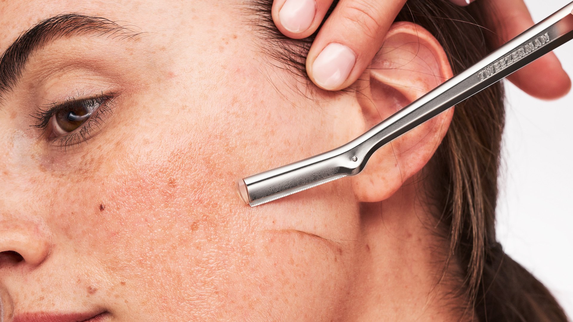 Model using a facial razor on her cheek with a silver facial razor tool.