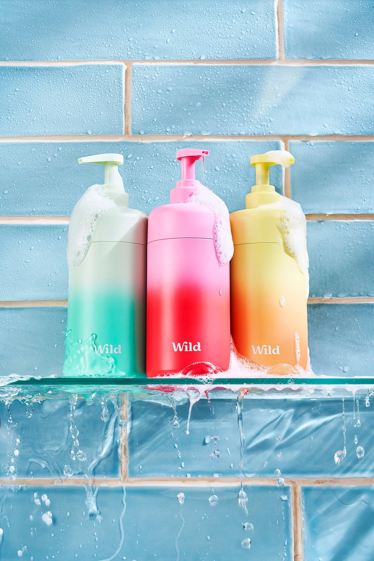 Three shower gel bottles are sitting on a glass shelf in a bathroom with blue tiles. Water splashing all around.