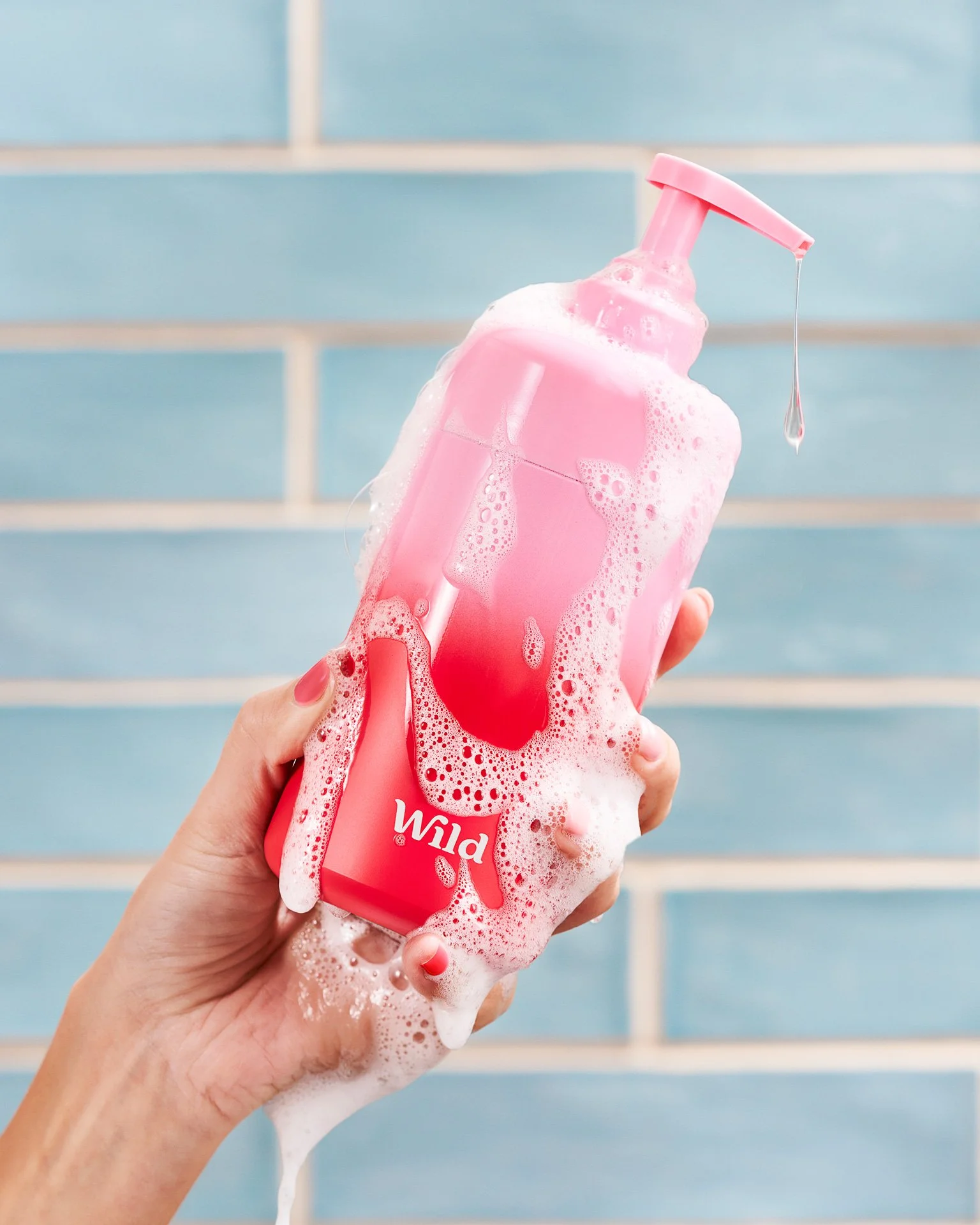 The model's hand is holding a pink and red ombre shower gel bottle, while covered in dripping suds.