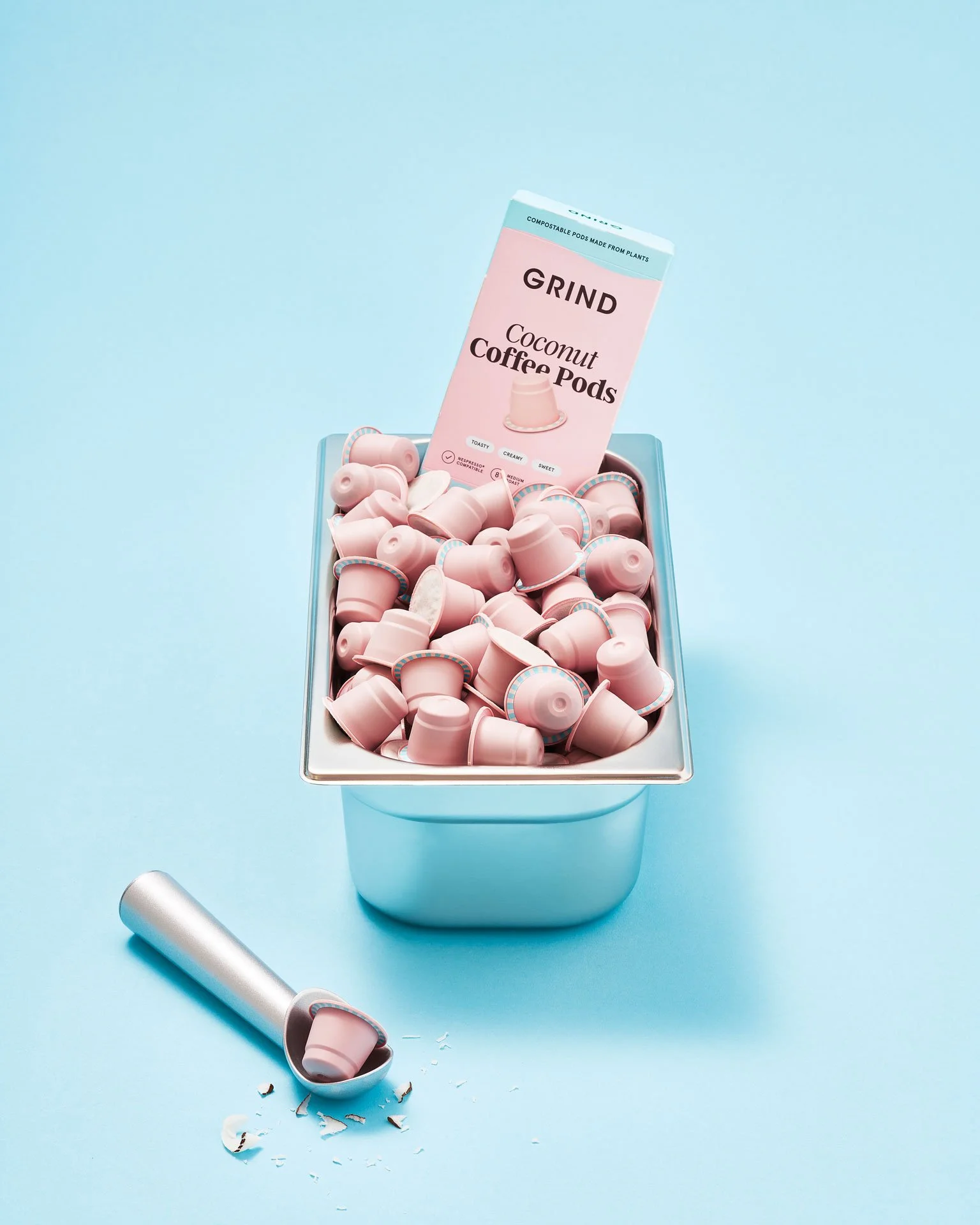 A metal container filled with pink coconut coffee pods, with one pod spilled out on the surface, some shards of the pod around it, and a cardboard box labeled "Grind Coconut Coffee Pods" standing among the pods.