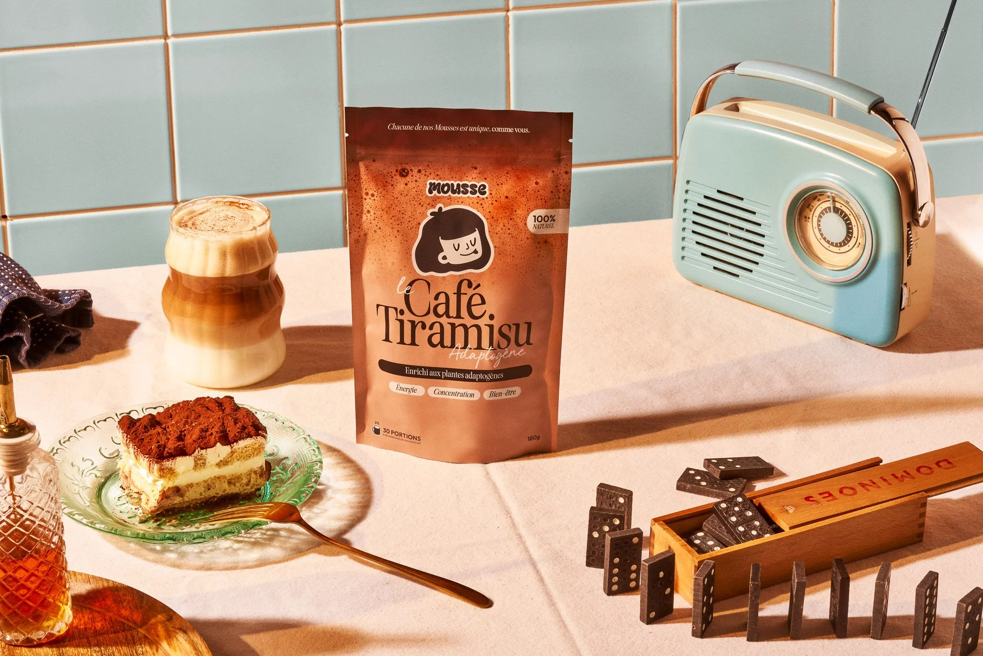 A tabletop with a slice of tiramisu cake on a glass plate, a fork, a mug with layered coffee, a package of mousse flavoring, an old-fashioned portable radio, and a dominoes game with domino tiles in a wooden box.