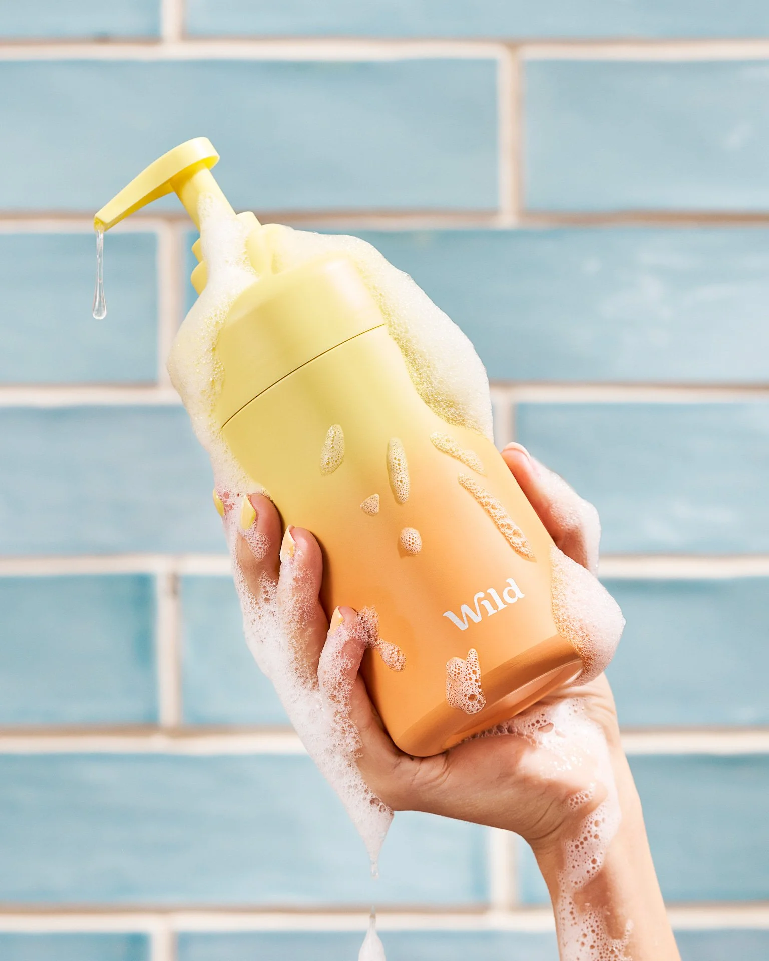 The model's hand is holding a yellow and orange ombre shower gel bottle, while covered in dripping suds.