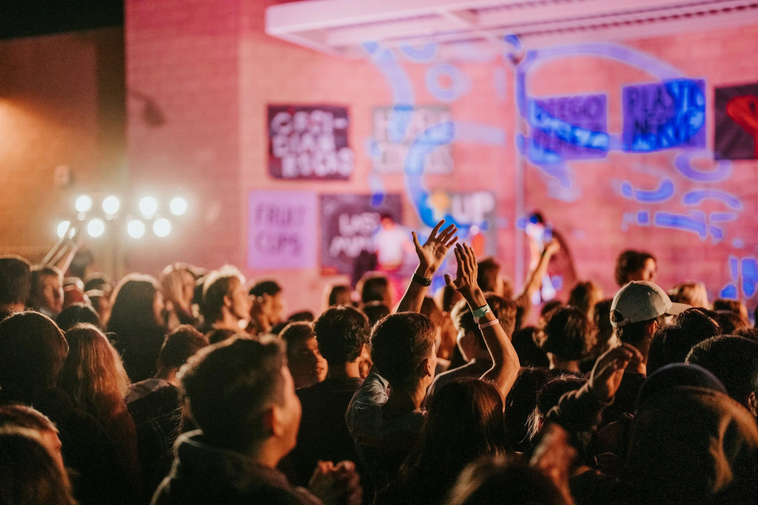 A large crowd of people gathered at an indoor concert or event, with some raising their hands. The background features a brick wall with various neon signs and lights, including words and symbols related to music and entertainment.