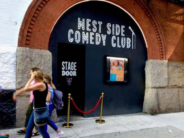 Entrance to West Side Comedy Club with a sign, stage door, and a poster display, red ropes, and people walking by.