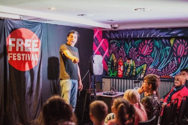 A young man on stage pointing at the camera during a stand-up comedy show at a free festival. The audience includes children and adults, with colorful murals and a fire extinguisher in the background.