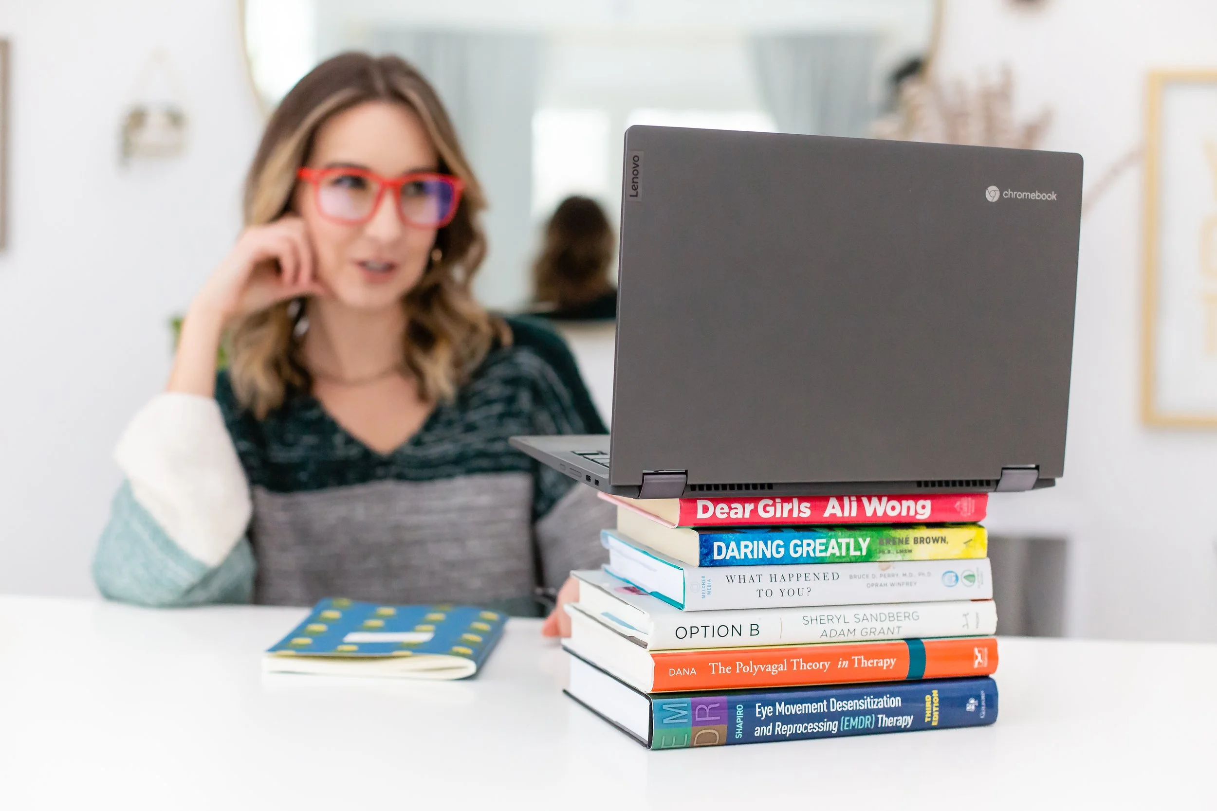 A woman with brown hair, red glasses, and a striped shirt sitting behind a desk with a stack of colorful books and a laptop with a Lenovo logo. The books include titles like "Dear Girls" by Ali Wong and "Eye Movement Desensitization and Reprocessing (EMDR) Therapy."