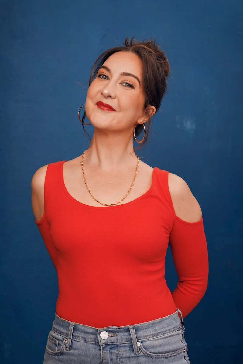 A woman with dark brown hair tied back, wearing a red top with cut-out shoulders, blue jeans, gold hoop earrings, a gold chain necklace, and red lipstick, standing against a blue background.