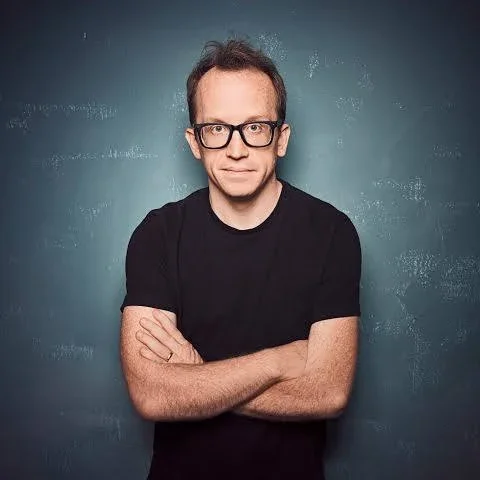 A man with glasses and short hair wearing a black T-shirt with crossed arms, standing against a textured blue background.