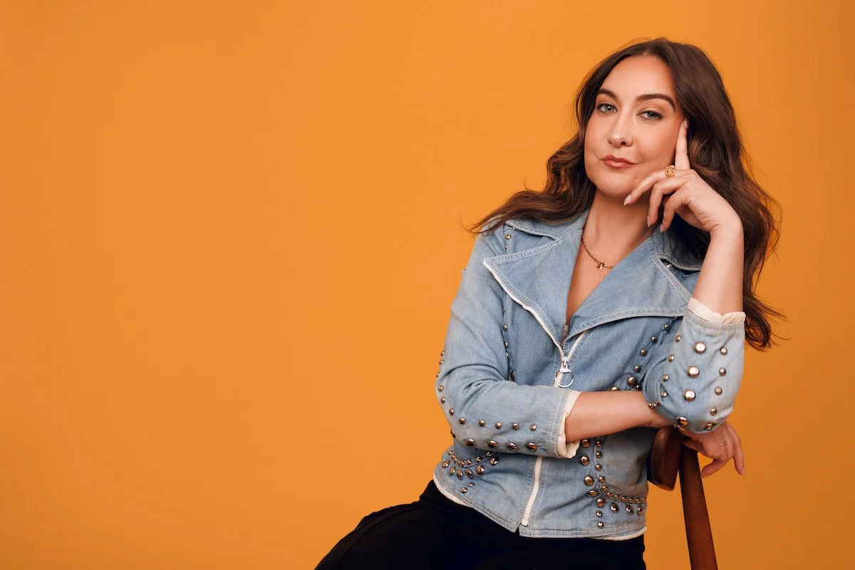 A woman with long wavy brown hair wearing a light blue denim jacket with metal studs, posing with her hand on her face, seated against an orange background.