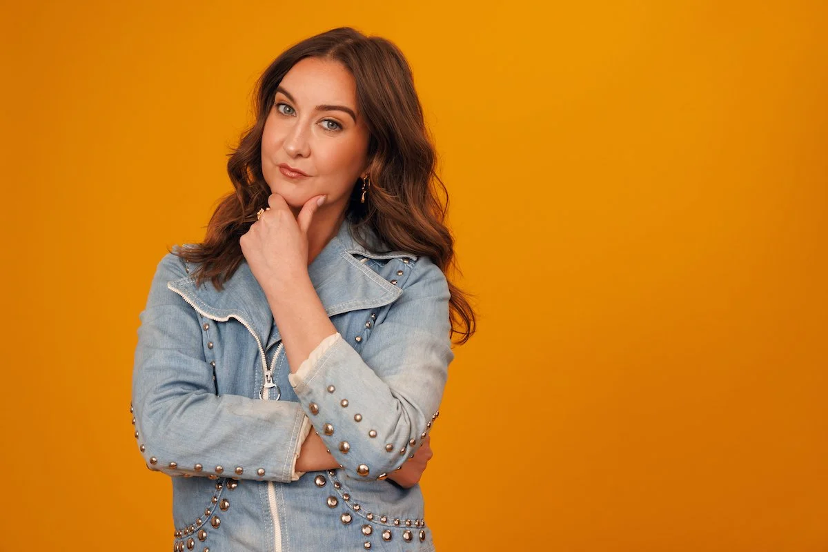 A woman with brown wavy hair, wearing a light blue denim jacket with metal stud embellishments, posing with her right hand resting on her chin against a solid orange background.